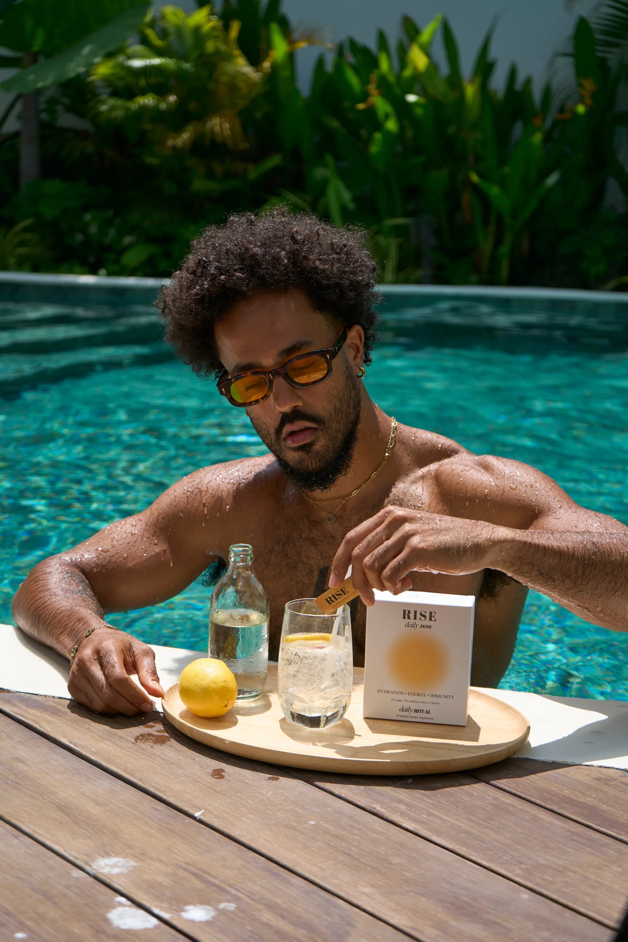 A man at the edge of a pool preparing a glass of RISE Daily Dose, the box beside him