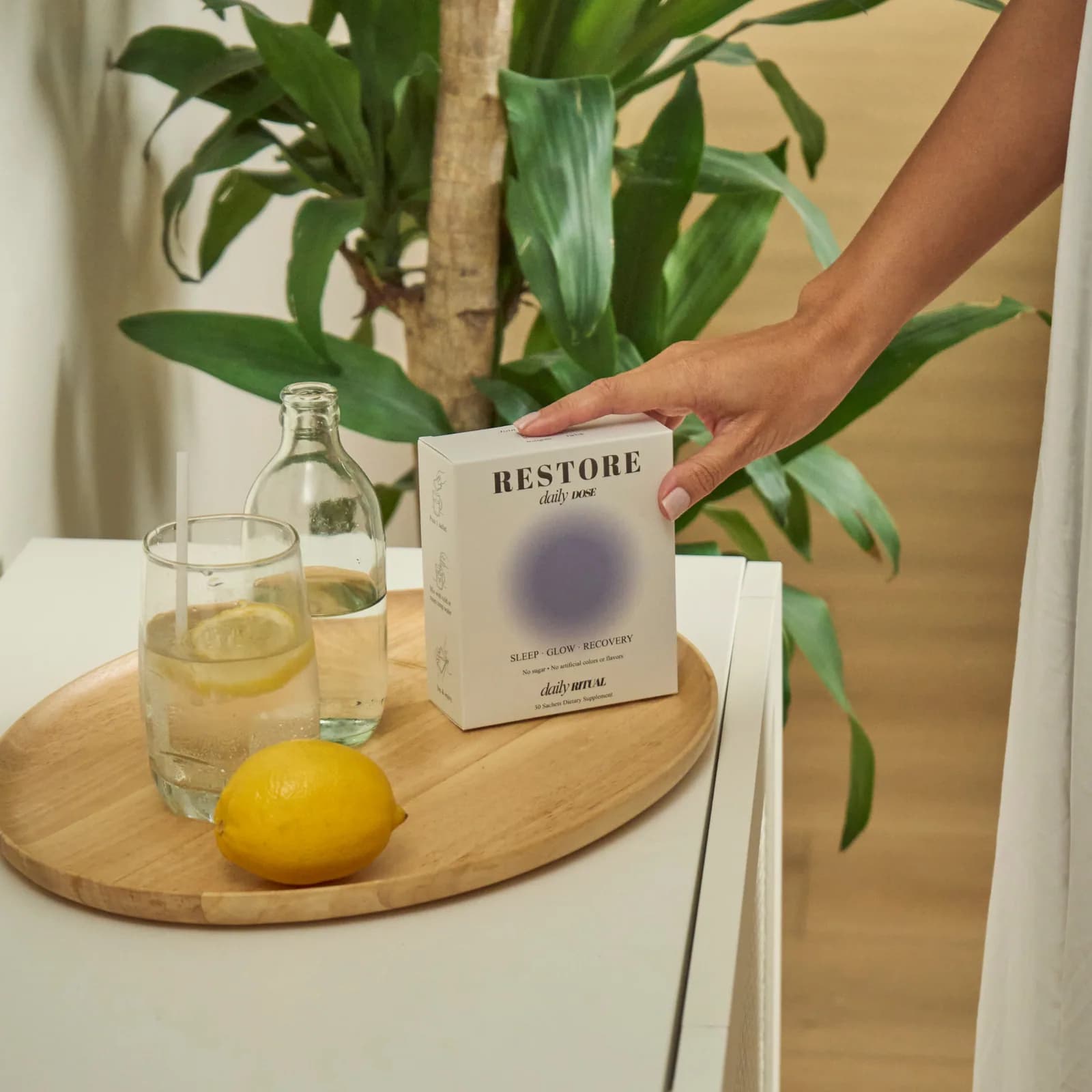 A hand setting down a box of RESTORE Daily Dose beside a glass of lemon water and a plant