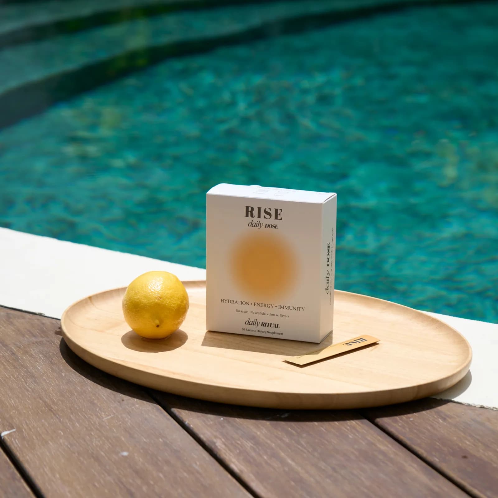 A box of RISE Daily Dose with a lemon on a wooden tray by the pool